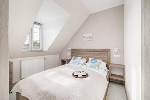 Un dormitorio con techo inclinado, una cama con cubrecama acolchado, dos mesitas de noche con lámparas, una ventana y una obra de arte colgada en la pared.