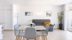 Un salon moderne avec une table à manger en verre, des chaises grises, un canapé gris agrémenté d’un coussin jaune et une grande plante en pot.