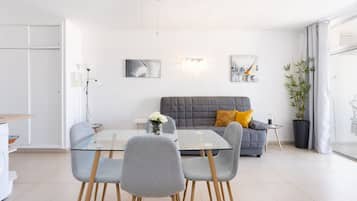 Una sala de estar moderna con una mesa de comedor de vidrio, sillas grises, un sofá gris con un cojín amarillo y una planta grande en maceta.