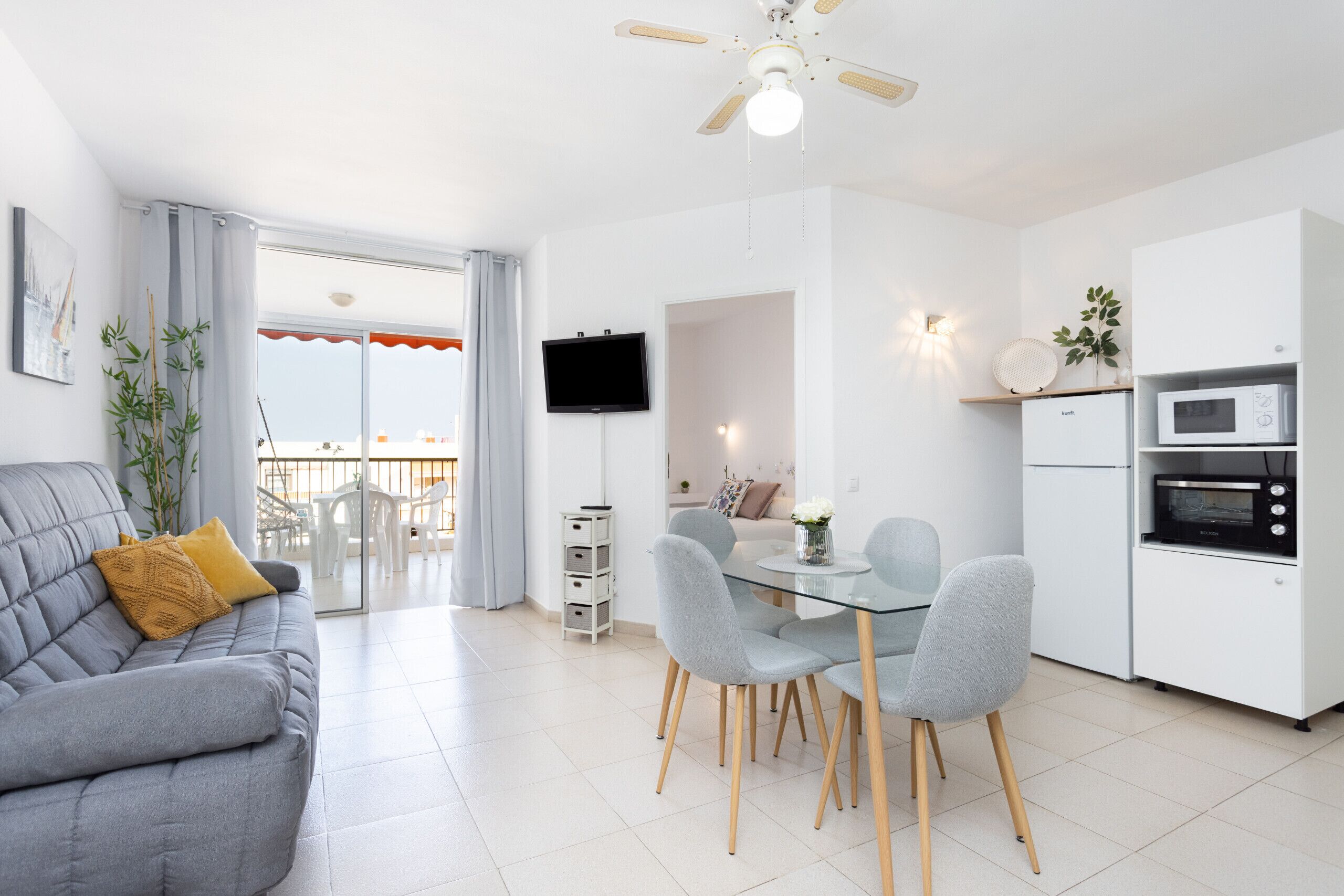 Una sala de estar moderna con un sofá gris, una mesa de comedor con sillas, una zona de cocina con armarios y electrodomésticos blancos, y un ventilador de techo.