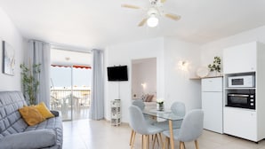 Un salon moderne avec un canapé gris, une table à manger avec des chaises, un coin cuisine doté de meubles blancs et d’appareils électroménagers, et un ventilateur de plafond.