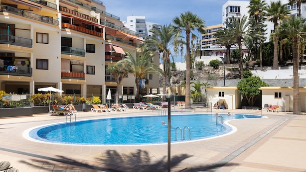 Une piscine entourée de chaises longues et de palmiers, avec des immeubles à plusieurs étages en arrière-plan.