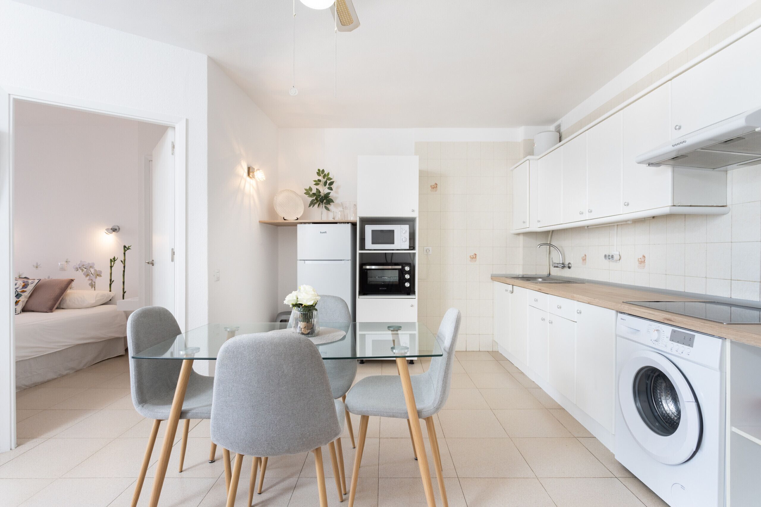 Een moderne keuken met een glazen eettafel, grijze stoelen, een wasmachine en een magnetron.