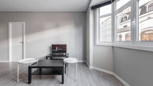 A modern living room with a dark wooden table, two white stools, a flat-screen TV, and large windows offering a view of buildings. - GuestReady - Olivieri's House - 7 (Porto)