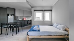 Une chambre moderne avec un lit en bois, un petit bureau et, au fond, un coin cuisine.