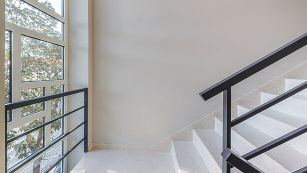 Un escalier avec une rampe, un mur blanc et une fenêtre donnant sur des arbres et un bâtiment.