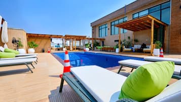 Un área de piscina en la azotea con sillones, una piscina y un edificio moderno con amplias ventanas.