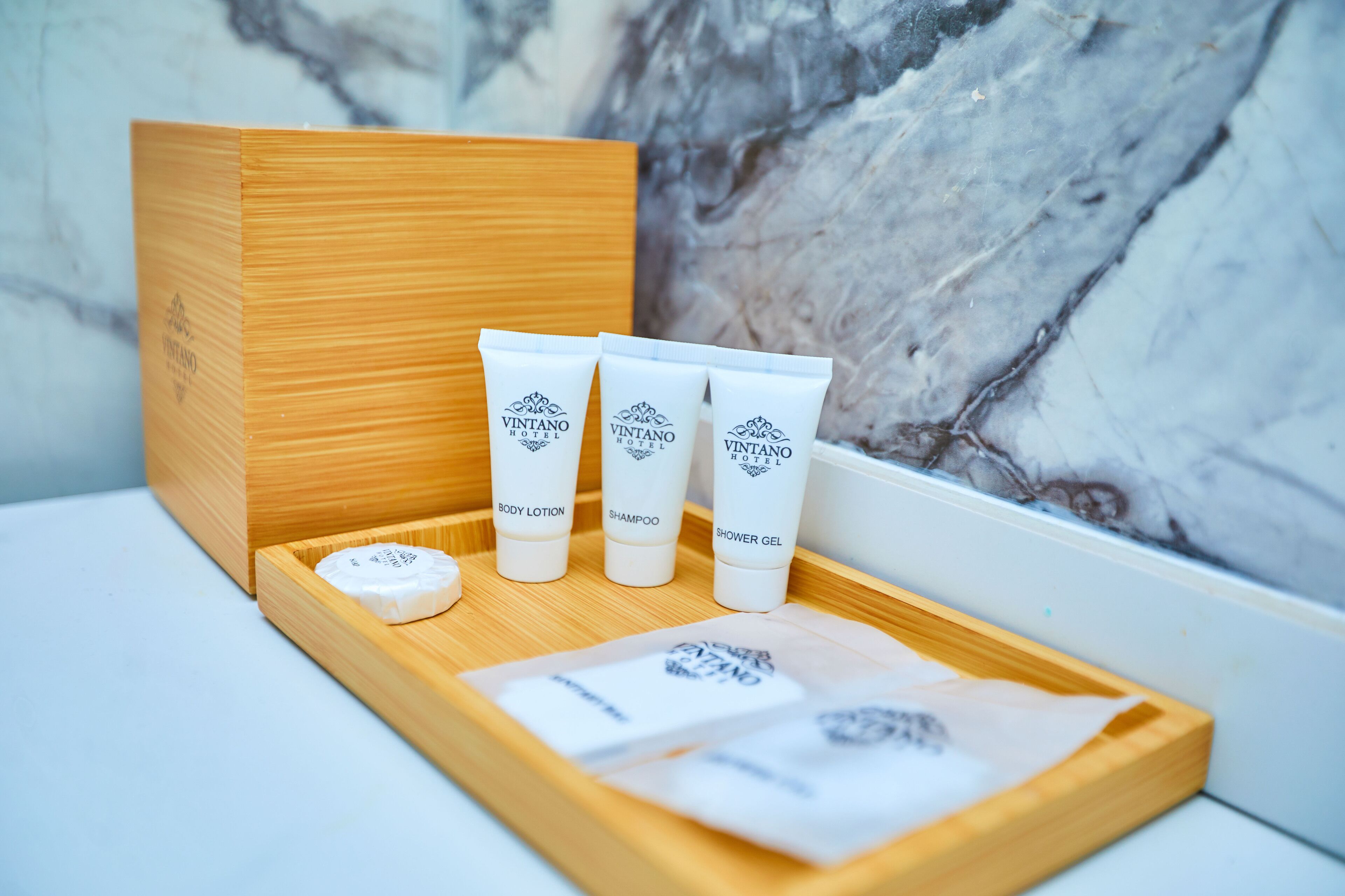 A wooden tray with three tubes of Vintano Hotel body lotion, shampoo, and shower gel, and a small round soap bar.