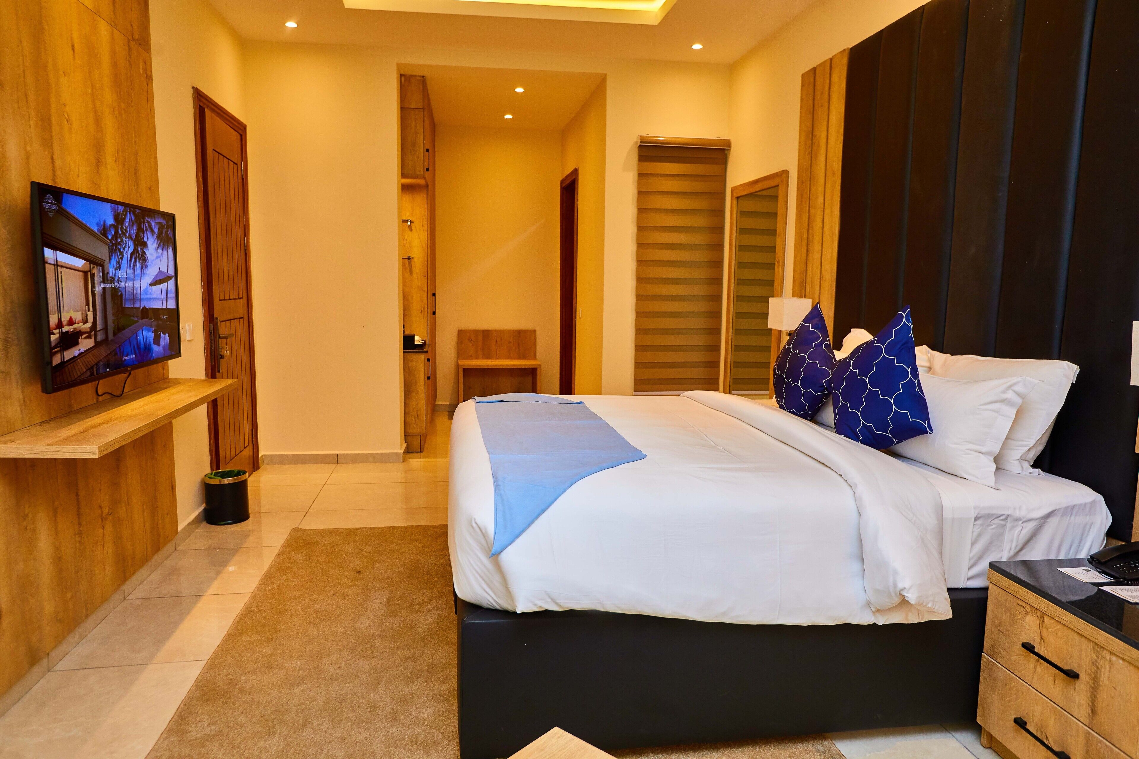 A modern hotel room with a large bed, a TV on the wall, wooden furniture, and a view of the hallway.