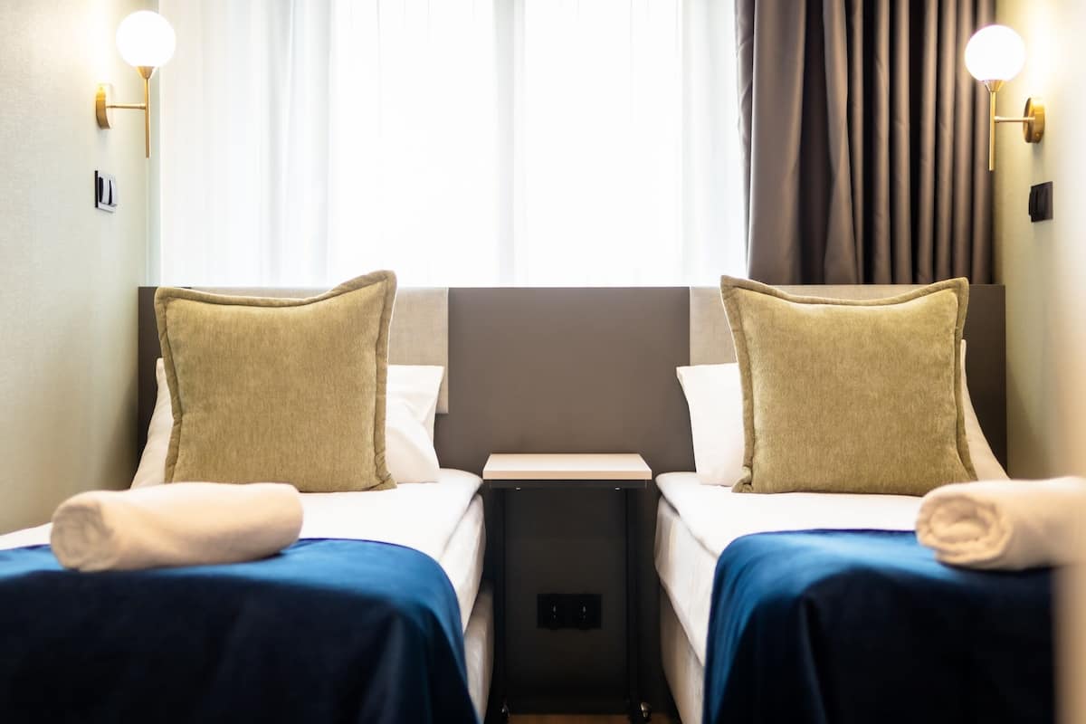 A hotel room with two beds, each with a pillow and a rolled towel. There are wall-mounted lights and a window with curtains.