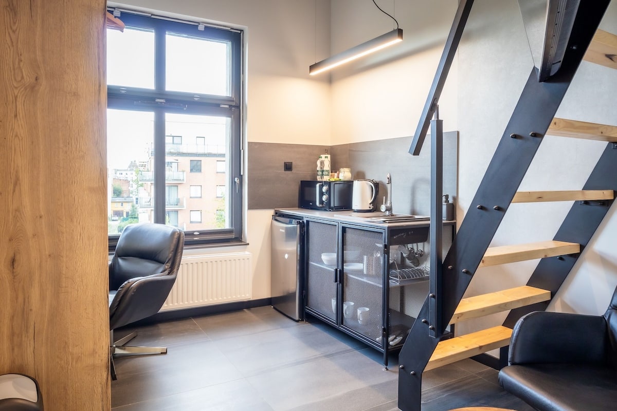 A compact kitchen with a built-in refrigerator, a microwave, and a coffee maker. There is a window providing natural light and a view of the outside. A staircase with wooden steps leads to an upper level.