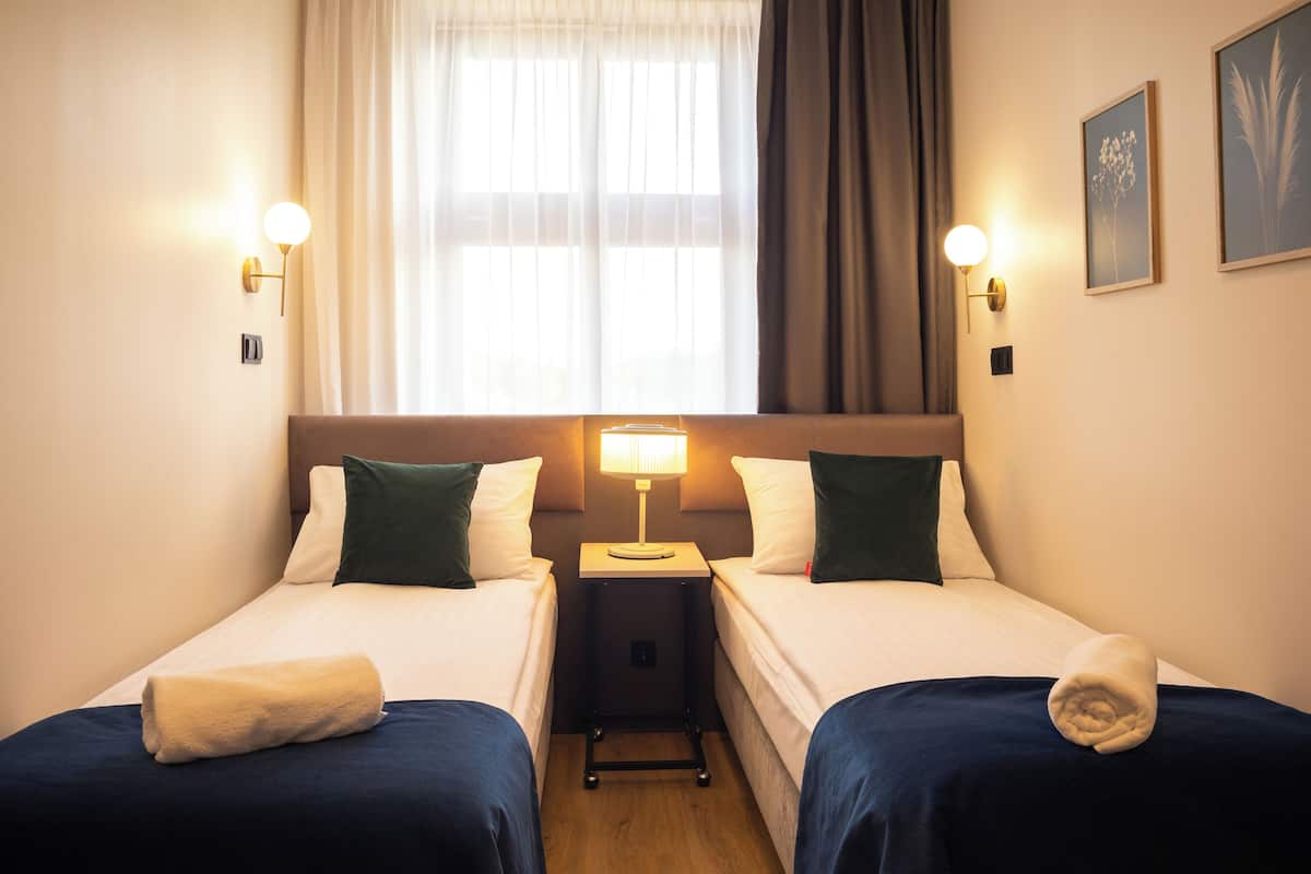 A hotel room with two single beds, a bedside table with a lamp, and a window with curtains.