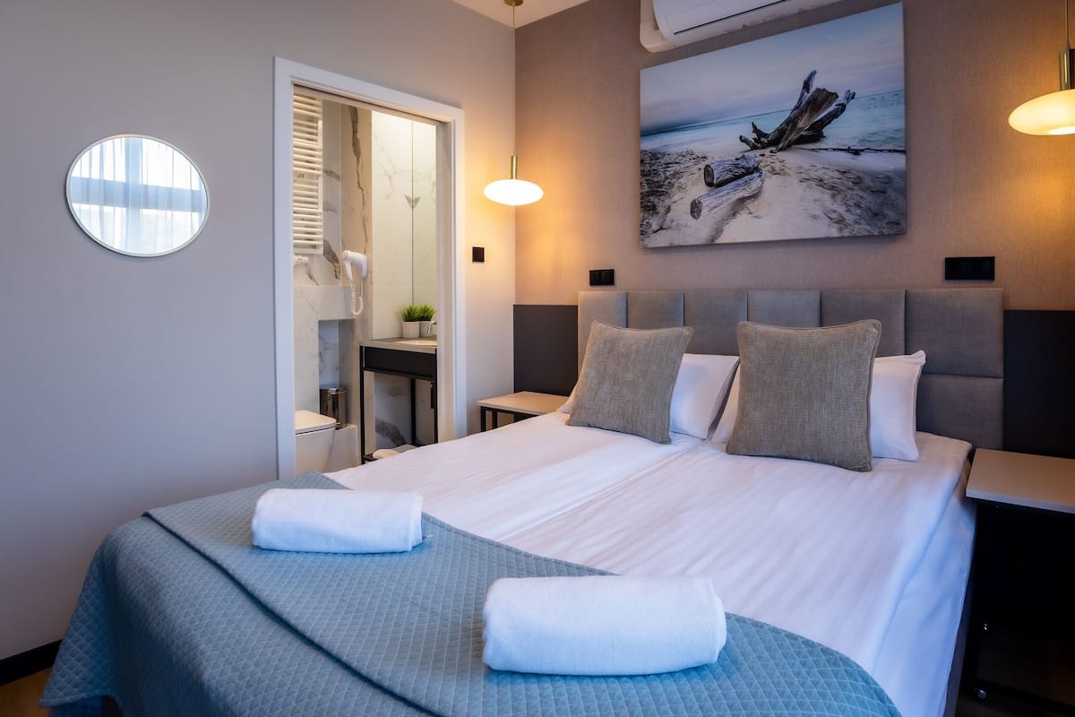 A neatly made bed with pillows, a bedside table, a round mirror, and a painting of a beach scene on the wall.