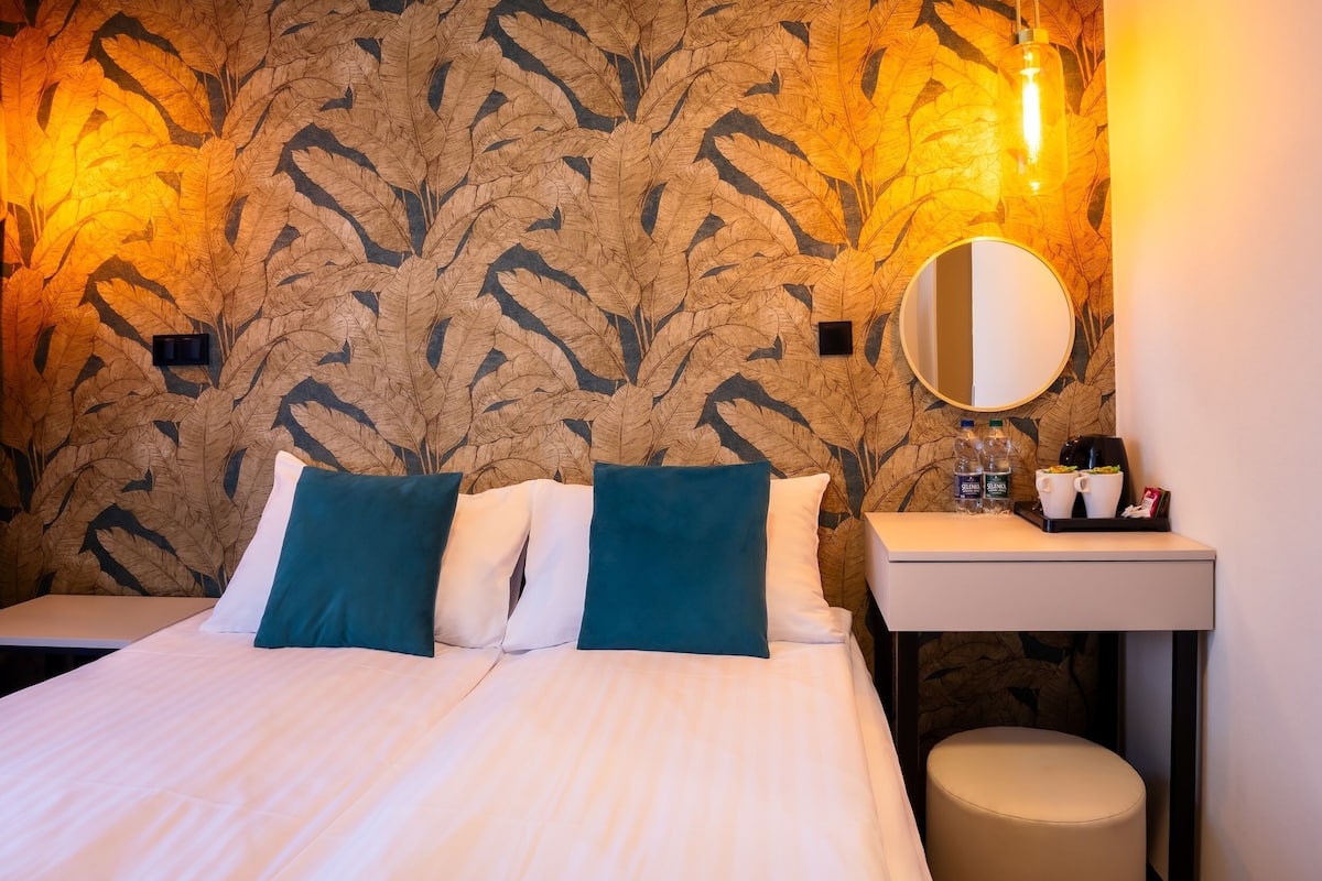 A hotel room with a bed, pillows, a bedside table with a mirror, and a small stool.