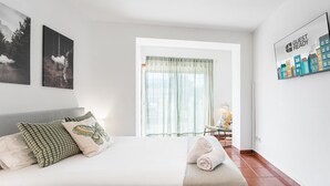 A bedroom with a large bed, a window with sheer curtains, a small table with a lamp, and artwork on the walls. - GuestReady - Portrait with a view (Porto)