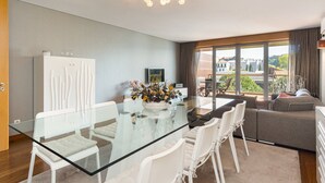 Ein modernes Wohnzimmer mit einem Glastisch, weißen Stühlen, einer Couch und einem großen Fenster mit Blick ins Freie.