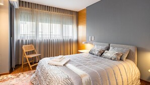 Ein modernes Schlafzimmer mit einem großen Bett, einem hölzernen Stuhl und einem Fenster mit transparenten Vorhängen.