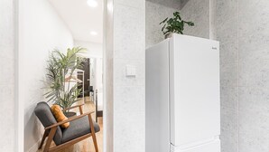 A modern kitchen with white tiled walls, a white refrigerator, and a wooden chair with a cushion. - GuestReady - Road to Revolution (Porto)