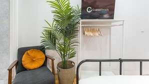 A neatly arranged room with a bed, a chair with a yellow pillow, a large potted plant, a wardrobe with wooden hangers, and a sign for GuestReady Group. - GuestReady - Road to Revolution (Porto)