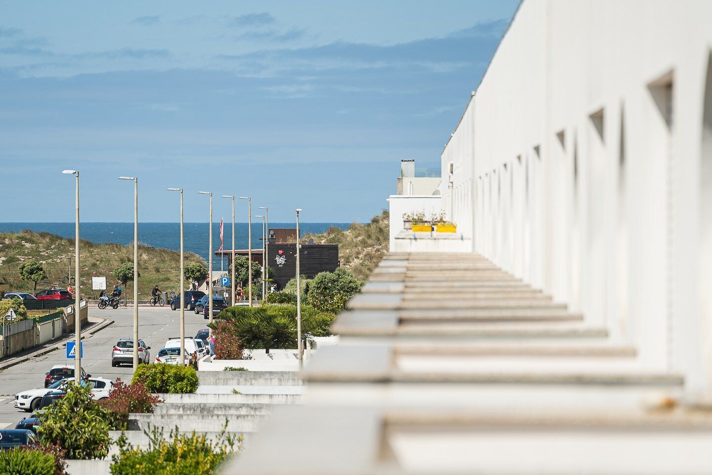 Uma estrada costeira com uma escadaria descendo até um edifício branco com janelas triangulares.