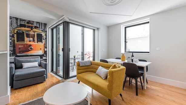 Un salon moderne avec un canapé, un fauteuil jaune, une table à manger et un coin cuisine visible à travers une porte vitrée.