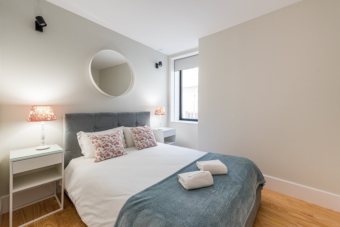 Une chambre moderne avec une tête de lit rembourrée de couleur grise, un miroir rond, deux tables de chevet avec des lampes, un lit avec une literie blanche et des coussins décoratifs, et une fenêtre qui laisse entrer la lumière naturelle.