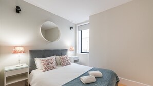 Une chambre moderne avec une tête de lit rembourrée de couleur grise, un miroir rond, deux tables de chevet avec des lampes, un lit avec une literie blanche et des coussins décoratifs, et une fenêtre qui laisse entrer la lumière naturelle.