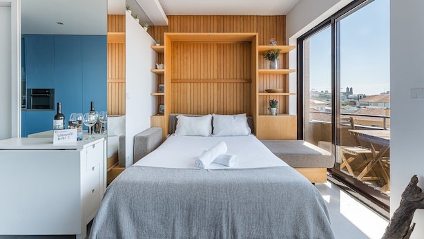 A modern bedroom with a large bed, a built-in bench, a wooden wall panel, a kitchen area, and a city view.