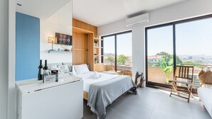 A modern hotel room with a bed, a desk with wine bottles, a balcony with a chair, and a city view.