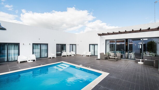 Un área moderna de piscina al aire libre con una piscina rectangular, sillones blancos para tomar el sol y un comedor con mesas y sillas.