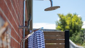 A showerhead attached to a metal wall, a towel hanging on a wooden bench, and a clear blue sky in the background. - Bundjil Beach House I Walk to beach (Bass Coast)