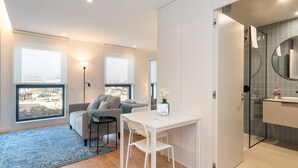 A modern hotel room with a sofa, a small table, a chair, a large window, and a bathroom with a glass shower enclosure. - GuestReady - A peaceful city refuge (Porto)