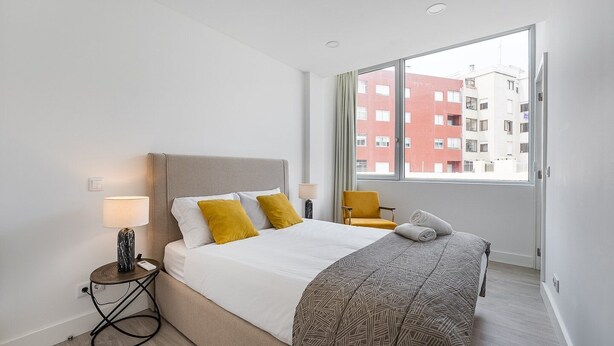 Quarto de hotel moderno com cama, mesa de cabeceira, abajur, cadeira e uma grande janela com vista para a cidade.