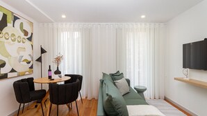 Een moderne woonkamer met een ronde houten tafel, twee zwarte stoelen, een groene bank met kussens, een witte vaas met bloemen en een schilderij aan de muur.