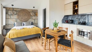 A modern bedroom with a stone wall, a bed with a yellow blanket, a dining table with chairs, and a kitchen area with a stove and microwave. - GuestReady - Caldeireiros Building (Porto)