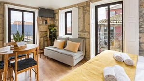 A room with a wooden dining table, a sofa with cushions, a bed with towels, and a TV mounted on a stone wall. - GuestReady - Caldeireiros Building (Porto)