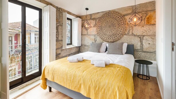A bedroom with a large bed, a stone wall, a round wall hanging, and a view of buildings outside. - GuestReady - Caldeireiros Building (Porto)