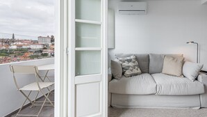 Una sala de estar moderna con un sofá, almohadas, una mesita y una silla. Hay una ventana con vistas a edificios y un paisaje urbano.