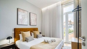 A modern hotel room with a large bed, a wooden headboard, two abstract wall art pieces, a bedside table with a lamp, and a view through a glass door to the outside. - GuestReady - Comendador Sto Ildefonso III (Porto)