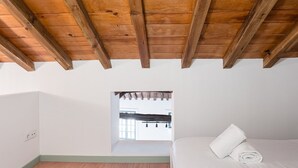 A bedroom with a sloped ceiling, wooden beams, a bed with white linens, and a view through a window to an outdoor area. - GuestReady - A brilliant stay near Ribeira (Porto)