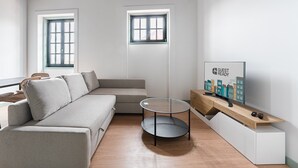 A modern living room with a sofa, a round glass coffee table, a TV on a wooden stand, and two windows with white frames. - GuestReady - A brilliant stay near Ribeira (Porto)