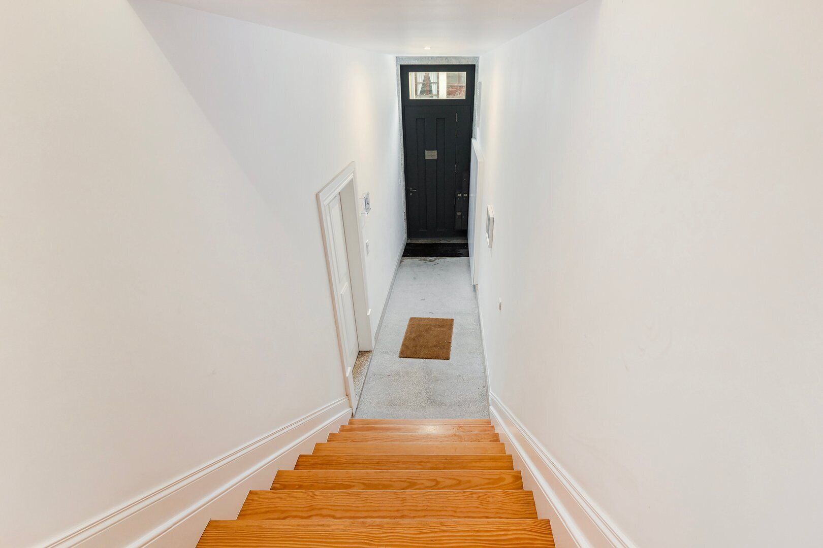 Un couloir étroit aux murs blancs, un escalier en bois et une porte noire au fond.
