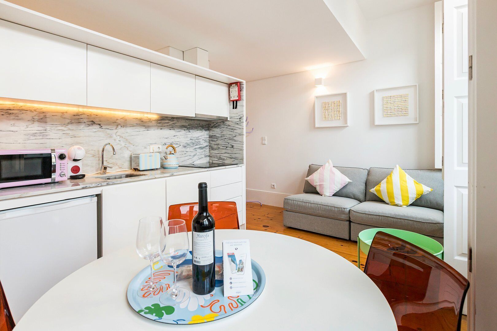 A modern kitchen with white cabinets, a marble countertop, and a pink microwave. There is a dining table with a wine bottle, glasses, and a placemat. A gray sofa with striped pillows is in the background.