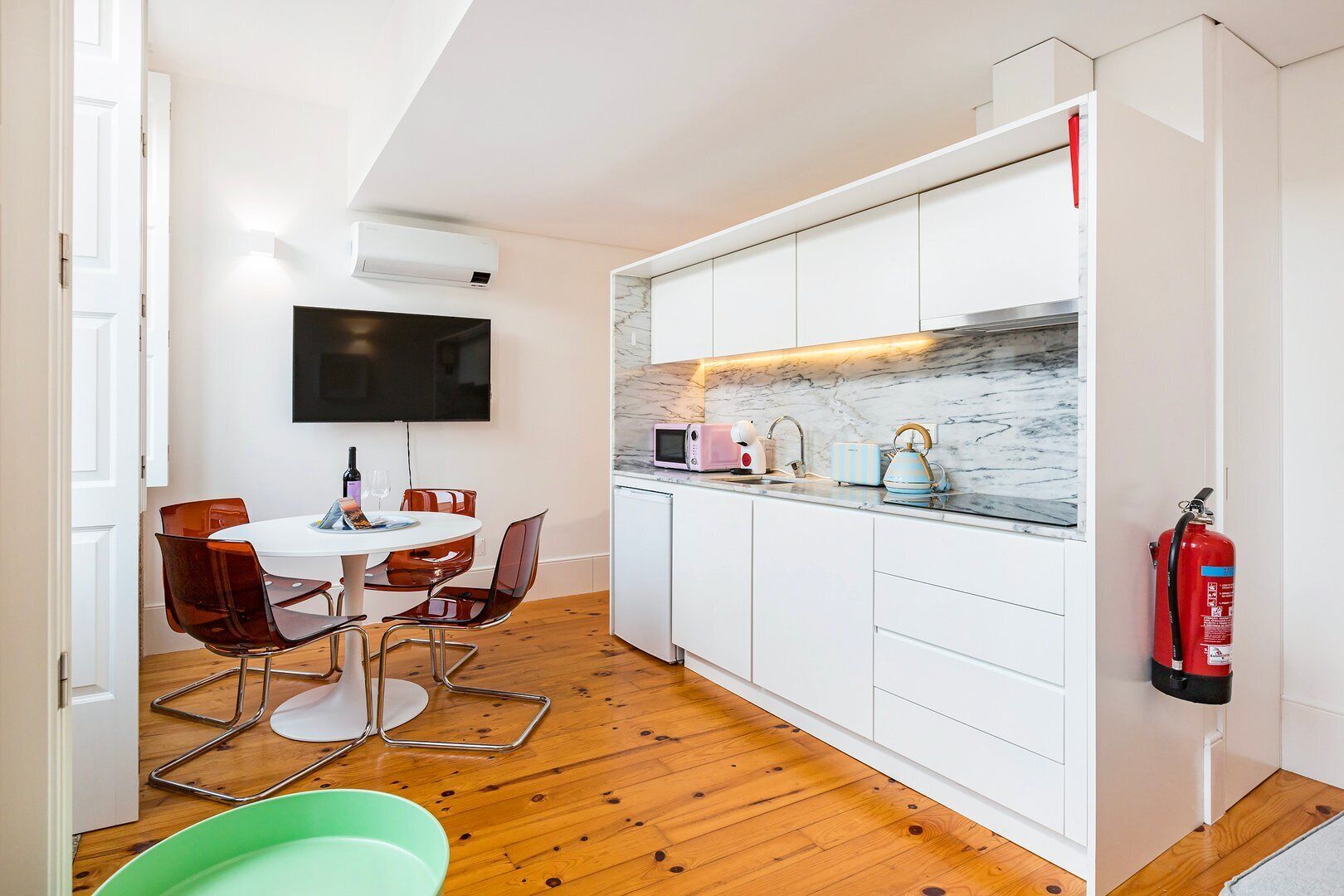 Une cuisine compacte avec des meubles blancs, un pare-feu en marbre, un petit coin repas avec une table ronde et des chaises, et un téléviseur à écran plat fixé au mur.
