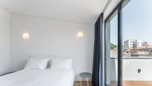 A modern bedroom with a large bed, a small side table, and a view of the city through a large window.