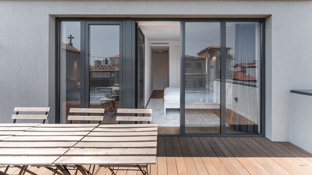 A modern balcony with a wooden table and chairs, a white sofa, and a glass door leading to an indoor space.