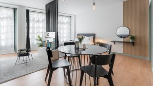 Et moderne hotelværelse med et rundt spisebord, sorte stole, en seng med gråt sengegavl, et stort vindue med lette gardiner, en væg af lodrette træpaneler og et rundt spejl, samt et skilt for GuestReady.