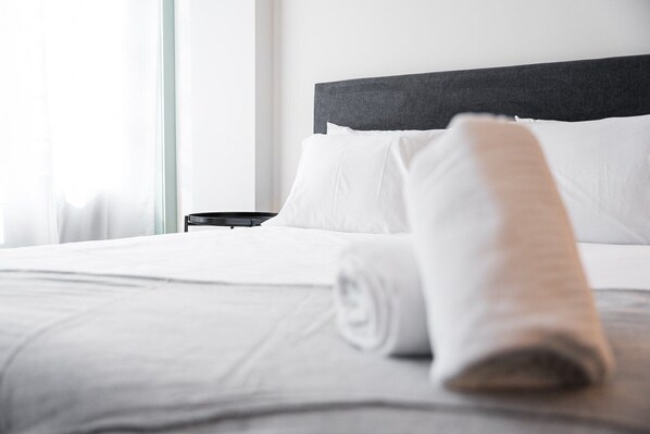 A neatly made bed with white linens and a rolled towel, a black headboard, and a small bedside table.