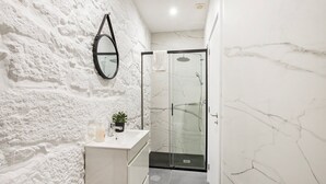 Une salle de bain moderne avec un mur en marbre blanc, un miroir rond, un petit lavabo agrémenté d’une plante et une douche à parois de verre.