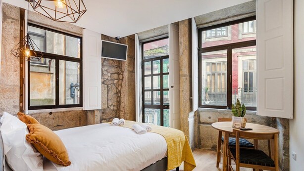 Une chambre à coucher avec un lit, une table et des chaises, de grandes fenêtres et un mur en pierre.